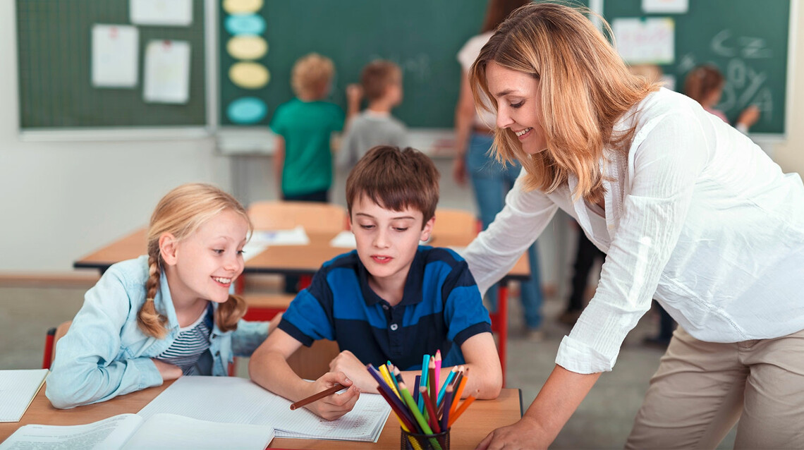 Lehrerin spricht mit zwei Grundschüler*innen im Klassenzimmer