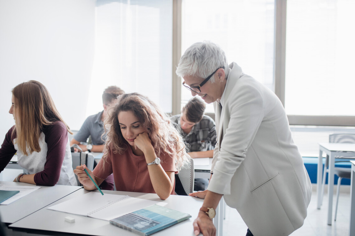Klassenlehrerin steht neben dem Tisch einer Schülerin und hilft ihr bei einer Aufgabe.