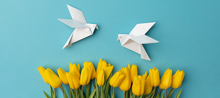 Zwei Origamivögel fliegen über gelben Tulpen vor einem blauen Hintergrund.