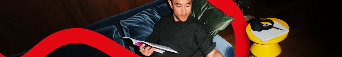 Junger Erwachsener liest auf Sofa im Lehrbuch und nimmt sich ein Stück Pizza, vorn schwebt ein illustriertes rotes Band