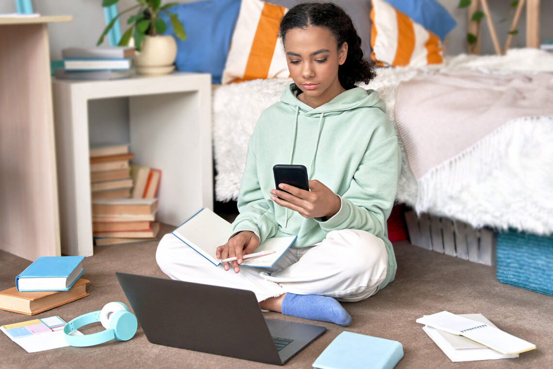 Schülerin sitzt mit Smartphone und ihrem Laptop auf dem Boden