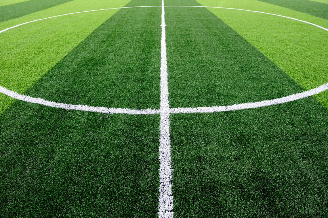 Fußballfeld mit Mittelkreis und weißen Linien auf grünem, gestreiftem Rasen.