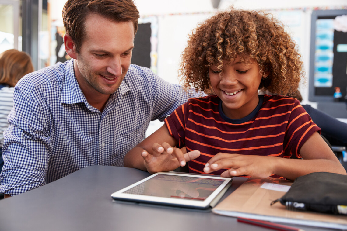 Lehrkraft sitzt neben einem Schüler vor einem Tablet