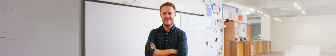 Eine junger, sympathischer Englischlehrer steht Klassenraum einer Berufsschule und lächelt selbstbewusst in die Kamera.