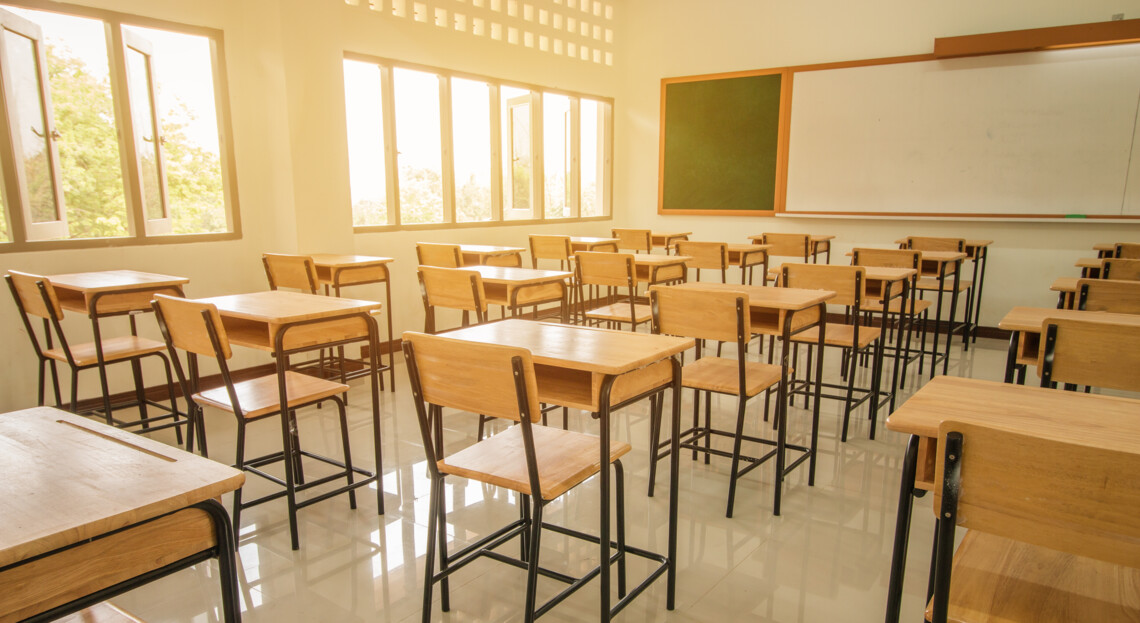 Ein leeres Klassenzimmer mit Holzstühlen und -tischen, Fenstern und einer grünen Tafel.