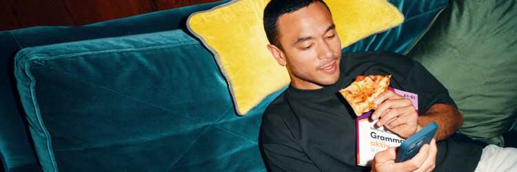 Junger Mann sitzt auf blauem Sofa mit gelbem Kissen, hält Pizza, Lehrwerk 'Grammatik aktiv' und Smartphone