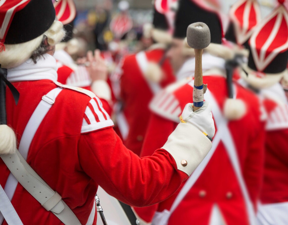 Eine Marschkapelle in roten Uniformen, ein Musiker schwingt einen Tambourstab.