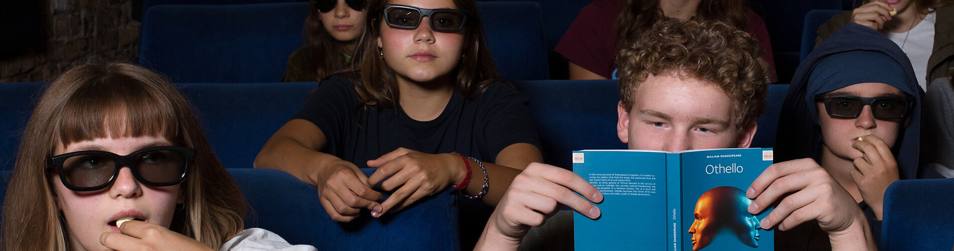 Menschen sitzen in einem Kino auf blauen Sitzen. Eine Person hält ein blaues Programmheft.